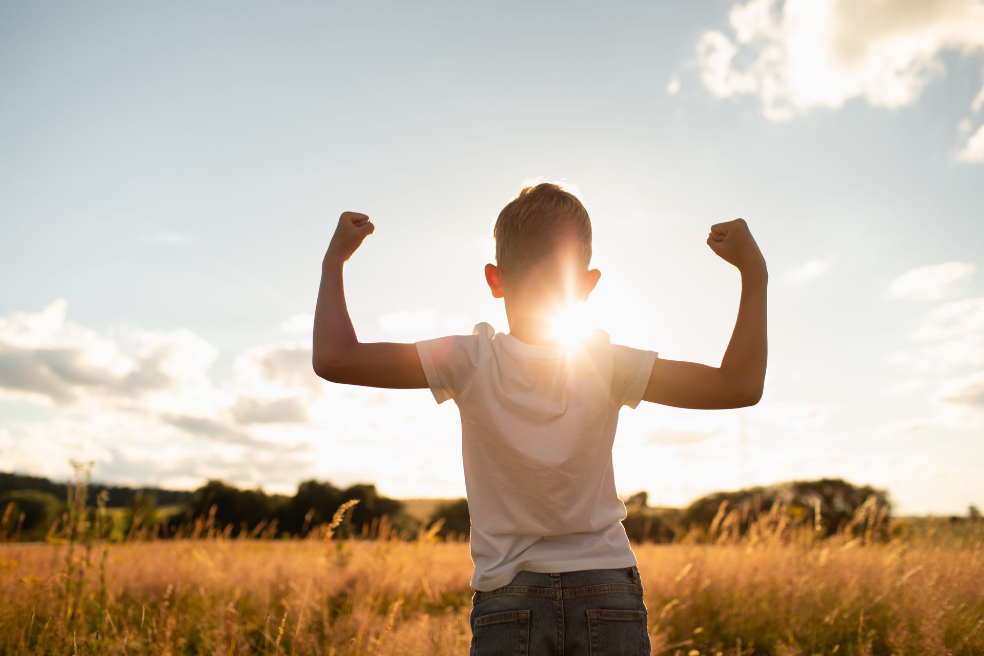 Un petit garçon fort fléchissant ses muscles dans la prairie au coucher du soleil.